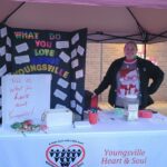 a heart & soul team member poses at a table they set up with a poster board that asks the question "what do you love about Youngsville?" Residents have used sticky notes to write their thoughts and attach them to the board.