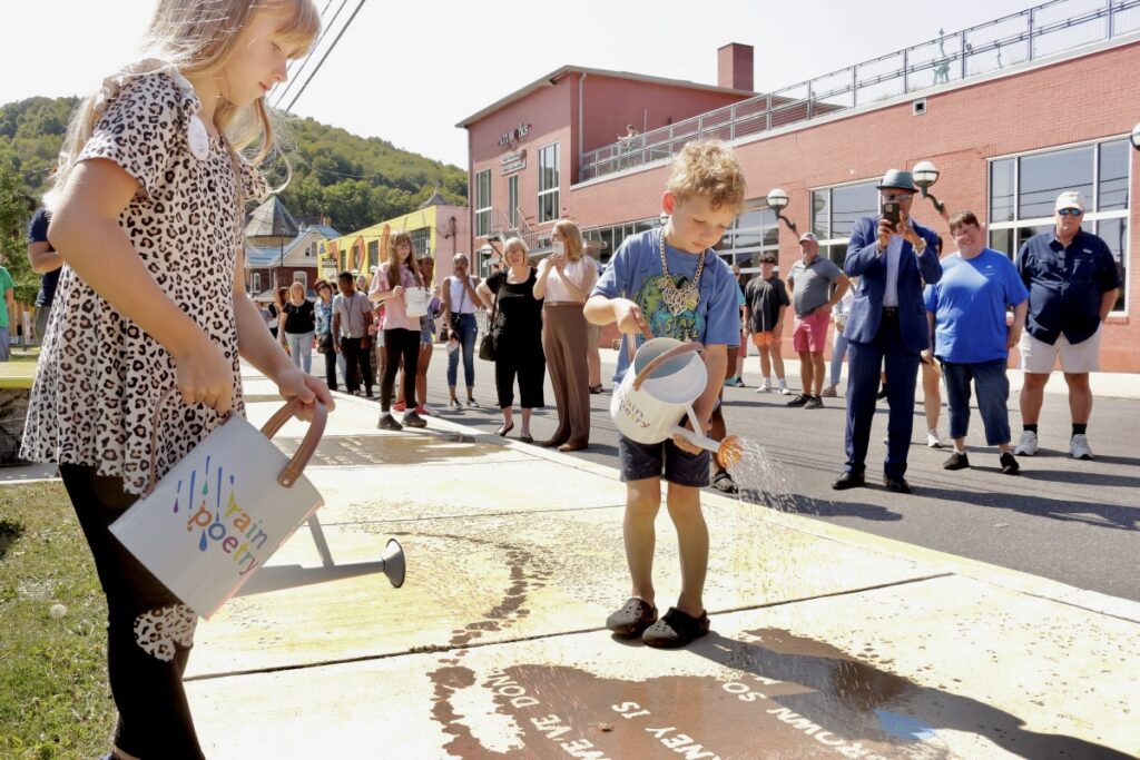 a young boy and girl use watering cans to sprinkle water on the sidewalk, making poetry appear like magic when the pavement gets wet