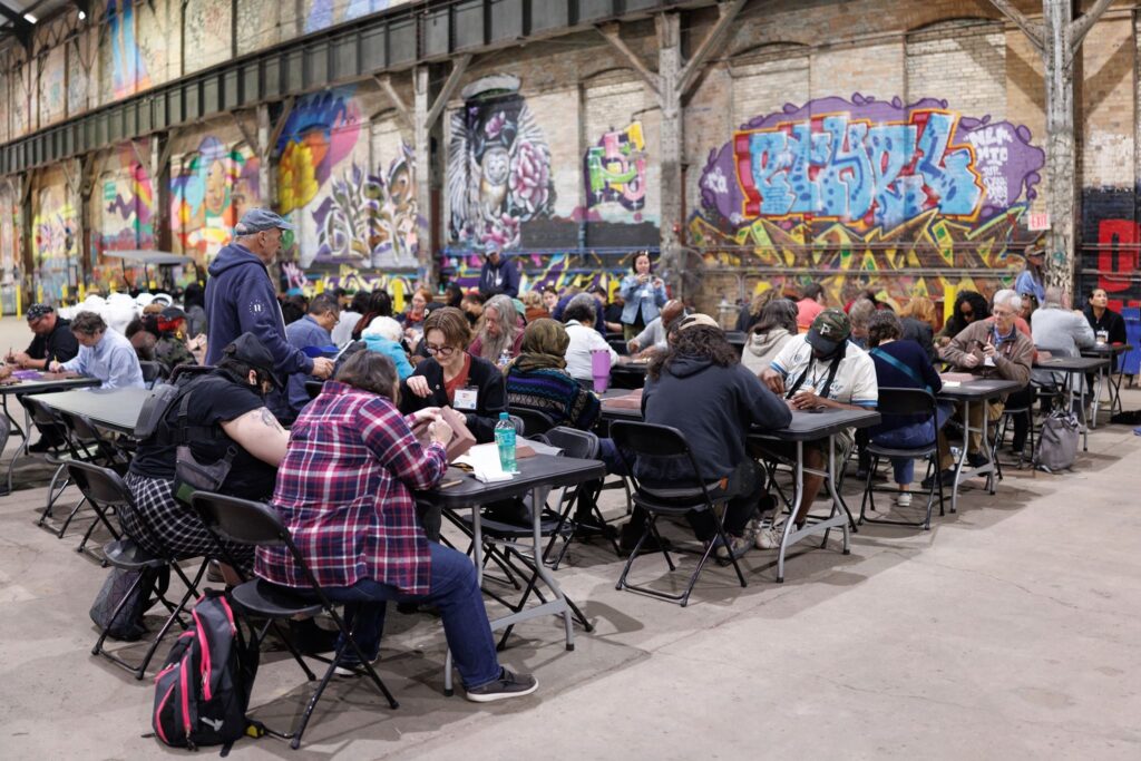 Fellows are seated at various long tables with graffiti art on the walls as they work on carving designs into molds to then cast in molten aluminum. 