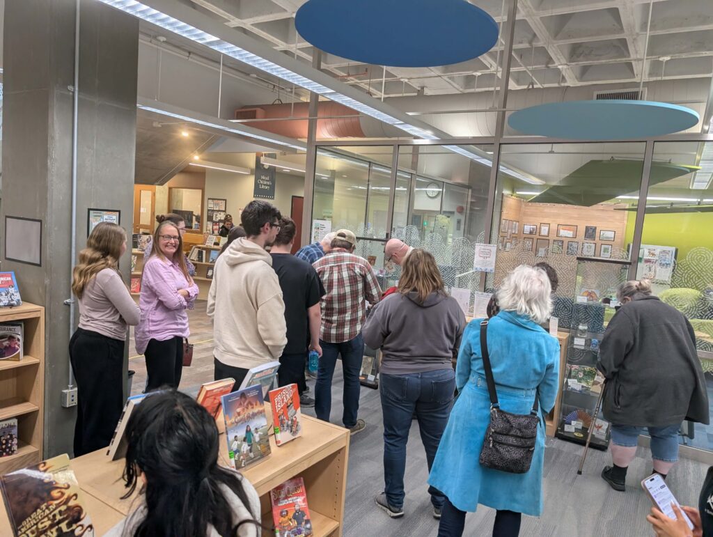 A group of approximately 15 adults are looking at display cases containing works created by teens during their exploration of folklore at the library.