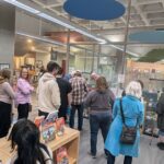 A group of approximately 15 adults are looking at display cases containing works created by teens during their exploration of folklore at the library.