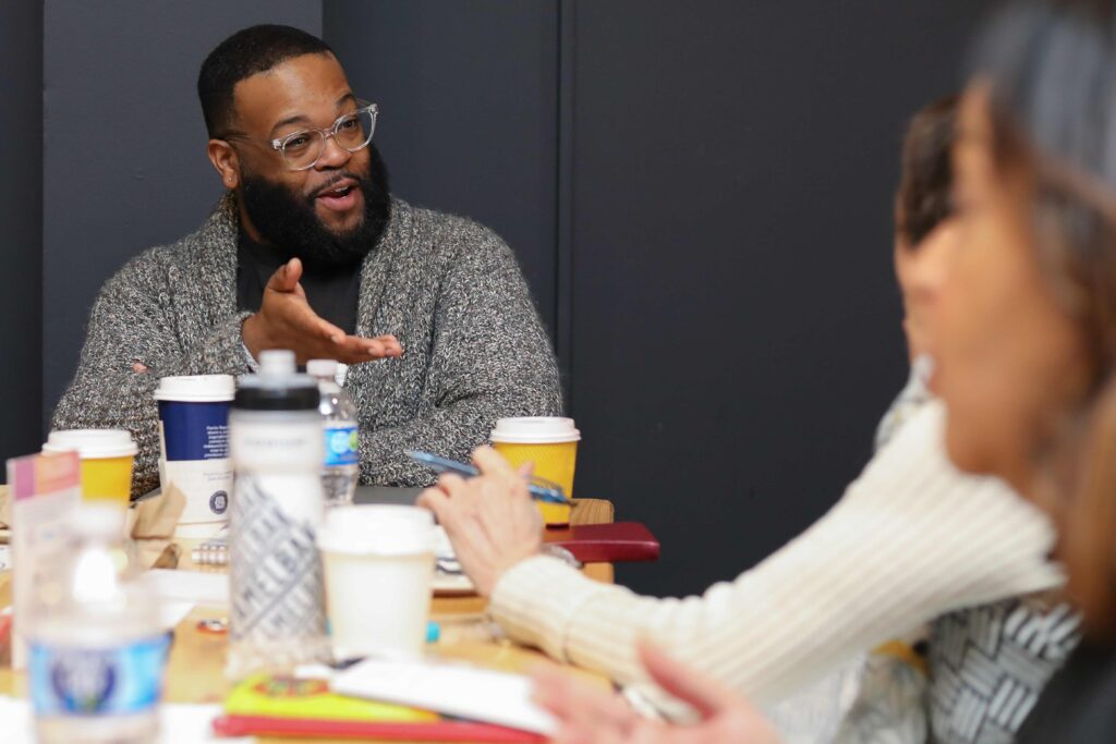 A man seated at a table gestures with his hand in conversation with a person who's shown only on the edge of the image.