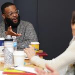 A man seated at a table gestures with his hand in conversation with a person who's shown only on the edge of the image.