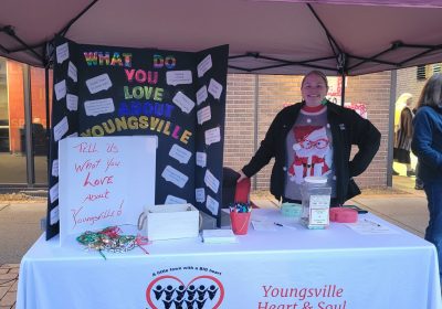a heart & soul team member poses at a table they set up with a poster board that asks the question "what do you love about Youngsville?" Residents have used sticky notes to write their thoughts and attach them to the board.