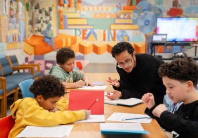 Teaching artist Adrian Perez-Roman leans over a table as he instructs three young students who are working on writing their poems.