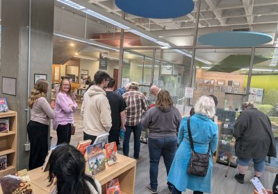 A group of approximately 15 adults are looking at display cases containing works created by teens during their exploration of folklore at the library.