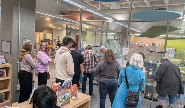 A group of approximately 15 adults are looking at display cases containing works created by teens during their exploration of folklore at the library.