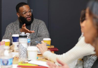 A man seated at a table gestures with his hand in conversation with a person who's shown only on the edge of the image.