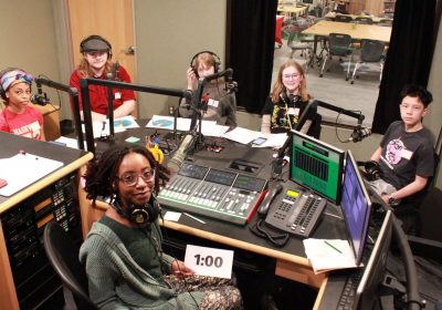five students and one educator are sitting in the radio booth smiling for the camera. the students are wearing headphones and each has a microphone in front of them. on the table that separates them sits equipment used for broadcasting.