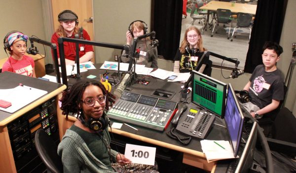 five students and one educator are sitting in the radio booth smiling for the camera. the students are wearing headphones and each has a microphone in front of them. on the table that separates them sits equipment used for broadcasting.