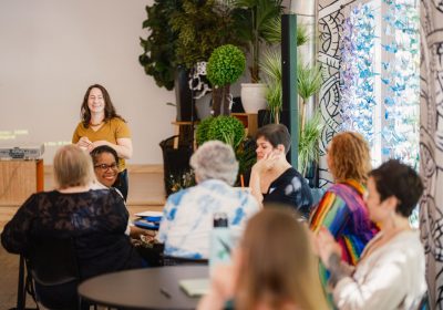 Julia Katz stands, smiling, wearing a mustard-colored top, at the front of the room in the background while a group of women who are attendees at the Youth-Led Humanities Community Culture Changers convening are seated around round tables, smiling and laughing.