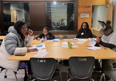 A group of four students ages 11 to 17 sit around a large table with papers spread in front of them. The student on the left, Dawn Martin, 17, is speaking. Behind them in the background is the studio booth where they'll conduct the radio show they're planning.