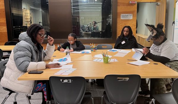 A group of four students ages 11 to 17 sit around a large table with papers spread in front of them. The student on the left, Dawn Martin, 17, is speaking. Behind them in the background is the studio booth where they'll conduct the radio show they're planning.