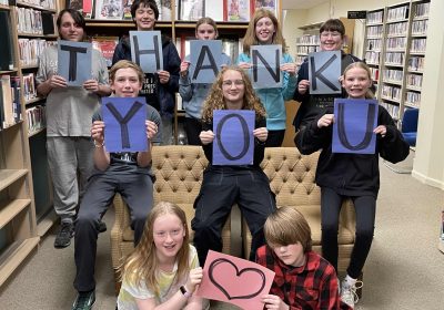 A group of teens at the library, some sitting on the ground in front, some leaning on a coach and others standing behind the couch, hold up lettered signs that spell out the words thank you.