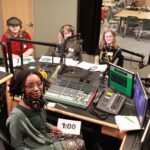 five students and one educator are sitting in the radio booth smiling for the camera. the students are wearing headphones and each has a microphone in front of them. on the table that separates them sits equipment used for broadcasting.