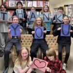 A group of teens at the library, some sitting on the ground in front, some leaning on a coach and others standing behind the couch, hold up lettered signs that spell out the words thank you.
