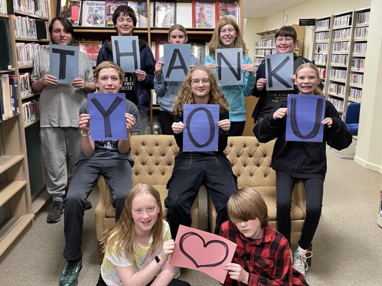 A group of teens at the library, some sitting on the ground in front, some leaning on a coach and others standing behind the couch, hold up lettered signs that spell out the words thank you.